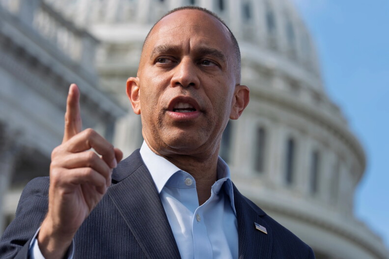 Hakeem Jeffries, who has medium-brown skin and closely cropped dark hair, gestures with a raised index finger while speaking, his gaze directed off to the side. He wears a navy pinstripe suit jacket with a small American flag lapel pin over a light blue open-collared shirt. The white dome of the U.S. Capitol is blurred behind him against a blue sky.
